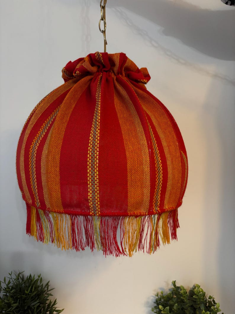 Peut inclure: Abat-jour suspendu avec un motif &agrave; rayures rouges et oranges. L'abat-jour a une forme ronde avec un dessus fronc&eacute; et une bordure &agrave; franges en bas. La frange comprend des fils rouges, jaunes et verts. L'abat-jour est suspendu par une cha&icirc;ne dor&eacute;e.