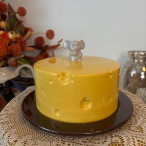 Peut inclure: Un plat à fromage en céramique jaune avec une base marron. Le plat à fromage présente des trous et une petite poignée grise en forme de souris sur le dessus. Le plat est posé sur une assiette marron, avec des décorations d'automne en arrière-plan.