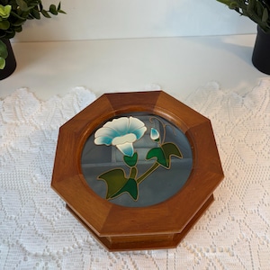 May include: Wooden octagon-shaped jewelry box with a glass top featuring a blue and white flower design. The box has a natural wood finish and a hinged lid. The flower design includes green leaves and stems.
