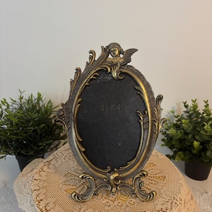 Può includere: Un'antica cornice ovale con una finitura in bronzo ornata. La cornice presenta un cherubino decorativo nella parte superiore e un'intricata voluta sui bordi. La cornice è supportata da una base decorativa ed è progettata per contenere una fotografia.