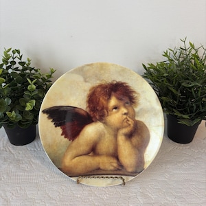 Könnte beinhalten: Ein dekorativer Teller mit einem Renaissance-Stil-Cherub mit rotem Haar und einem einzelnen Flügel. Der Cherub ist mit einem nachdenklichen Ausdruck dargestellt, der sein Kinn auf seine Hand stützt. Der Teller wird auf einem goldfarbenen Metallständer präsentiert.
