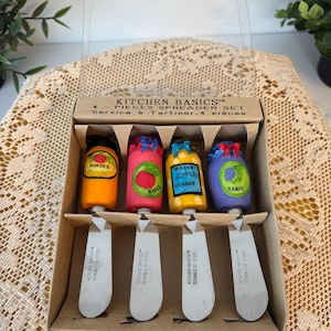 Peut inclure: Un ensemble de quatre couteaux à tartiner Kitchen Basics dans une boîte en carton. Chaque couteau a une lame en acier inoxydable et un manche coloré avec un design de fruit différent : orange, pomme, citron et raisin. La boîte indique "Service à Tartiner. 4 pièces."