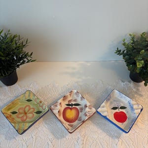 Peut inclure: Trois coupelles carrées en céramique aux coins arrondis, chacune ornée de motifs peints à la main. La coupelle de gauche présente une fleur rose, celle du centre une pomme jaune et rouge, et celle de droite une pomme rouge aux bords bleus.
