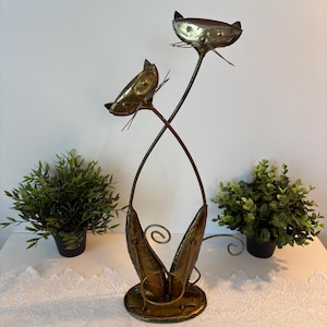 Peut inclure: Une sculpture vintage en métal représentant des chats avec deux têtes sur de fines tiges. La sculpture a une finition bronze et une base circulaire. Les visages des chats sont détaillés avec des moustaches. Deux petites plantes en pot flanquent la sculpture.