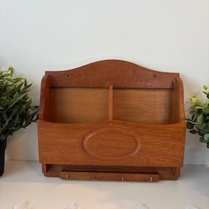 Puede incluir: Un organizador de pared de madera con un acabado marrón cálido. Cuenta con dos compartimentos, un diseño ovalado y una fila de ganchos debajo. El organizador está diseñado para el almacenamiento de correo y llaves.