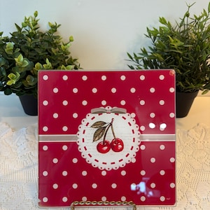 Puede incluir: Plato cuadrado rojo con lunares blancos y diseño de cerezas. El plato presenta un círculo festoneado blanco con una ilustración de cerezas y una cinta plateada. Un artículo decorativo para la cocina, de unos 20 cm x 20 cm.