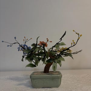 May include: A small bonsai tree ornament in a rectangular, pale green ceramic pot. The tree has a brown trunk, green leaves, and colourful blossoms in blue, orange, and yellow. The bonsai is a decorative item.