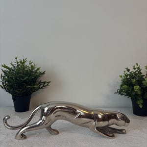 May include: A silver-colored, decorative panther statue in a crouching pose. The statue has a sleek, reflective surface and is placed on a white lace tablecloth. Two small potted plants with green foliage flank the statue.