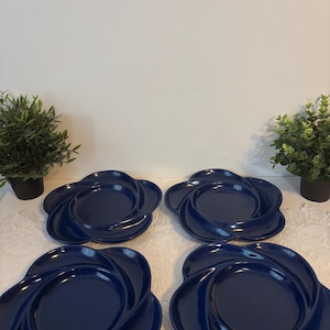 Könnte beinhalten: Vier marineblaue, blütenförmige Teller. Jeder Teller hat eine zentrale ovale Fläche, die von wirbelnden, blütenblattartigen Rändern umgeben ist. Die Teller sind auf einer weißen Oberfläche angeordnet, mit kleinen Topfpflanzen im Hintergrund.