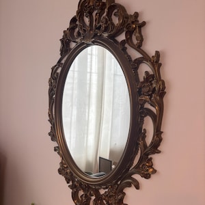May include: An oval mirror with an elaborate, bronze-coloured frame. The frame has floral and scrollwork details. The mirror is hanging on a pink wall, reflecting a room with a window and curtains.