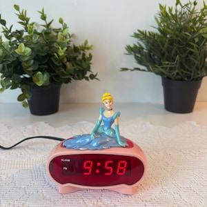 May include: A pink digital alarm clock featuring a Cinderella figurine. The clock shows the time 9:58 in red numerals. The alarm clock has an alarm function and is connected to a power cable.