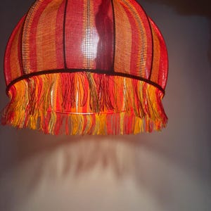 Peut inclure: Un abat-jour vintage aux couleurs vives. L'abat-jour pr&eacute;sente des rayures verticales dans les tons de rouge, d'orange et de jaune. Le bord inf&eacute;rieur est orn&eacute; d'une frange de fils jaunes et oranges. L'abat-jour est suspendu &agrave; une cha&icirc;ne.