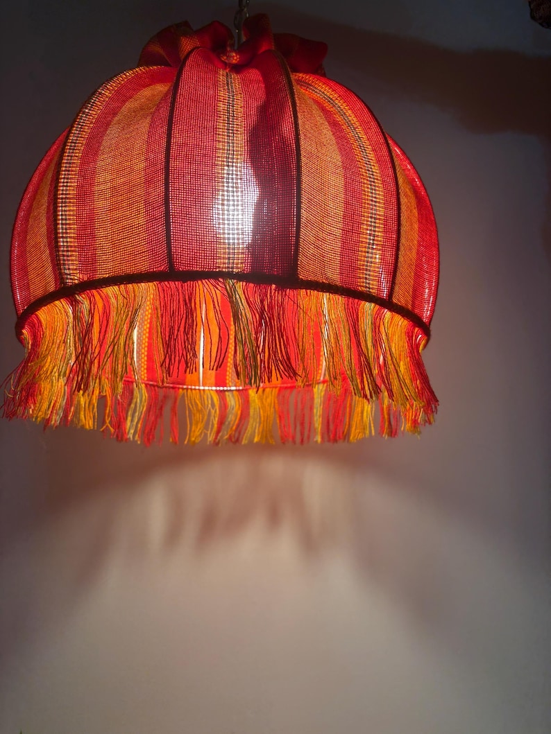 Peut inclure: Un abat-jour vintage aux couleurs vives. L'abat-jour pr&eacute;sente des rayures verticales dans les tons de rouge, d'orange et de jaune. Le bord inf&eacute;rieur est orn&eacute; d'une frange de fils jaunes et oranges. L'abat-jour est suspendu &agrave; une cha&icirc;ne.