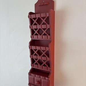 May include: A wall-mounted wooden organiser painted in a deep matte red. The unit features two open storage compartments with a lattice-style front design and a single small drawer at the bottom with a round knob, all finished in a solid red colour.