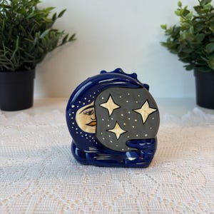 May include: A ceramic figurine of a crescent moon with a face peeking out from behind a grey cloud with four gold stars. The moon is dark blue with white dots. The figurine is on a white lace tablecloth.
