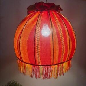 Peut inclure: Un abat-jour suspendu, de forme sph&eacute;rique, avec des rayures rouges et oranges. Il est orn&eacute; d'une frange en bas et d'un volant en haut. La lumi&egrave;re chaude de l'ampoule est visible &agrave; travers le tissu.