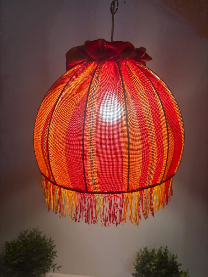 Peut inclure: Un abat-jour suspendu, de forme sph&eacute;rique, avec des rayures rouges et oranges. Il est orn&eacute; d'une frange en bas et d'un volant en haut. La lumi&egrave;re chaude de l'ampoule est visible &agrave; travers le tissu.