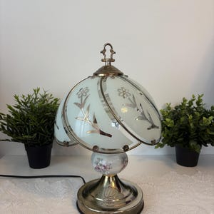 Peut inclure: Une lampe de table vintage avec un motif floral. La lampe présente un abat-jour en verre bleu clair avec des accents floraux argentés et une base en laiton. La base a un motif floral et une base ronde de couleur laiton. La lampe mesure environ 30 cm de haut.