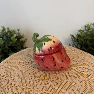 Peut inclure: Une boîte à bijoux en céramique en forme de fraise avec un couvercle. La boîte est principalement rouge avec des mouchetures noires et une tige et des feuilles vertes. Le couvercle s'adapte parfaitement à la base.