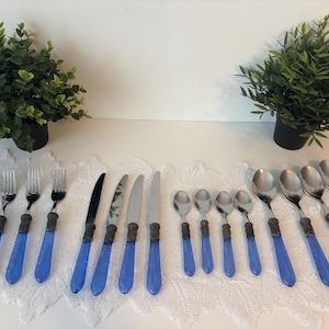 Puede incluir: Un juego de cubiertos vintage con mangos azules. El juego incluye tenedores, cuchillos y cucharas. Los mangos son de color azul vibrante y las partes metálicas tienen un acabado plateado. Los cubiertos están dispuestos sobre un mantel de encaje blanco.