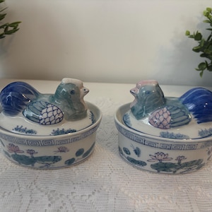 Könnte beinhalten: Zwei keramische Schmuckdosen in Entenform. Die Enten haben blaue, grüne und rosa Details. Die Dosen sind weiß mit blauen Blumen- und geometrischen Mustern. Die Deckel sind wie Enten geformt und die Körper sind rund.