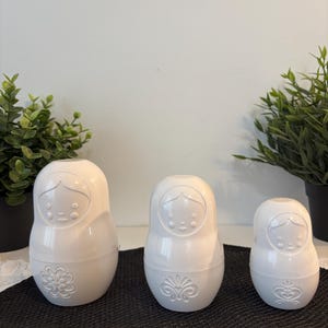 Peut inclure: Trois poupées russes blanches de tailles différentes. Chaque poupée présente un visage stylisé et un motif floral ou ornemental décoratif sur la partie inférieure. Les poupées sont disposées sur une surface noire avec de la verdure en arrière-plan.
