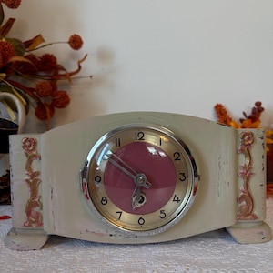 May include: Cream-coloured vintage mantel clock with a pink clock face and silver-tone trim. The clock has decorative pink accents on the sides and a curved top. The clock is on a white lace tablecloth.