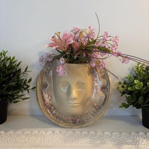 Peut inclure: Une jardinière murale en céramique beige en forme de visage humain, dotée d'un bord circulaire avec des motifs de plumes en relief. La jardinière est remplie d'un arrangement de lys roses artificiels et de petites fleurs violettes décoratives.