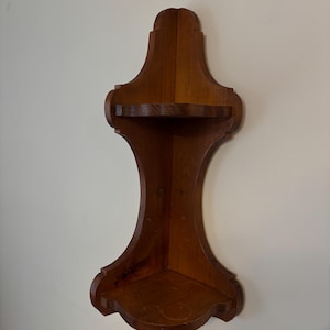 May include: A wooden corner shelf with a dark brown finish. The shelf has two tiers and a decorative, curved design. It is mounted on a white wall.