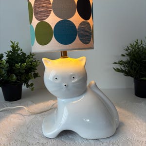 May include: A white ceramic cat-shaped table lamp with a patterned lampshade. The lampshade features large circles in shades of green, blue, gray, and black. The lamp is on a white surface, with small potted plants on either side.