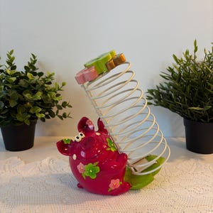 Peut inclure: Une figurine de cochon magenta vif avec des motifs floraux verts et roses, fixée à un support en spirale en fil métallique blanc. La spirale est surmontée d'une pièce en forme de fleur verte et rose, sur une base en forme de feuille verte.