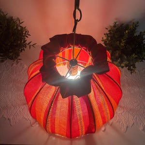 Peut inclure: Une lampe suspendue avec un abat-jour en tissu ray&eacute; rouge et orange. L'abat-jour a un dessus &agrave; volants et est suspendu par une cha&icirc;ne. La lampe est allum&eacute;e, d&eacute;gageant une lueur chaleureuse.