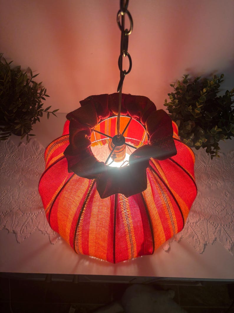 Peut inclure: Une lampe suspendue avec un abat-jour en tissu ray&eacute; rouge et orange. L'abat-jour a un dessus &agrave; volants et est suspendu par une cha&icirc;ne. La lampe est allum&eacute;e, d&eacute;gageant une lueur chaleureuse.