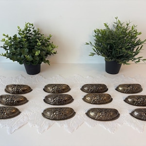 Peut inclure: Ensemble de poignées de tiroir antiques de couleur bronze avec un motif floral. Les poignées sont disposées sur une surface blanche, avec deux plantes vertes en pot en arrière-plan. Idéal pour la restauration de meubles.