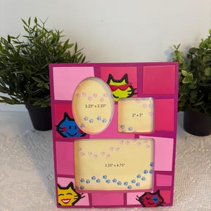 May include: A pink and magenta multi-photo frame with cat-themed designs. The frame has three photo openings: an oval (3.25" x 2.25"), a square (2" x 2"), and a rectangle (3.25" x 4.75"). Each opening is decorated with paw prints.