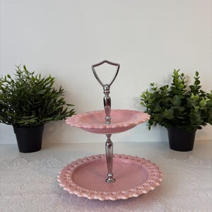 以下が含まれることがあります： シルバーのハンドルが付いた、2段のピンク色のサービングトレイ。トレイはスカラップエッジのデザインです。ハンドルは、シールド型のトップが付いたシルバーメタルです。トレイは白いレースのテーブルクロスの上に展示されています。