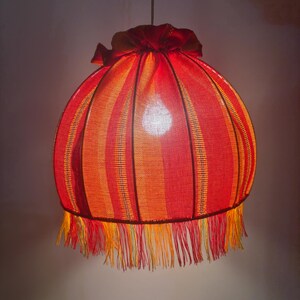 Peut inclure: Suspension luminaire avec abat-jour ray&eacute; rouge et orange. L'abat-jour est de forme arrondie, avec une frange en bas et un sommet fronc&eacute;. La lampe est suspendue &agrave; une cha&icirc;ne.