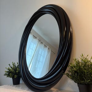 Peut inclure: Un miroir ovale doté d'un cadre noir brillant au design torsadé et tourbillonnant. Le cadre se compose de plusieurs bandes arrondies superposées qui créent un effet de spirale continu autour de la surface réfléchissante centrale en verre.