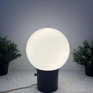 Peut inclure: Une lampe de table composée d'un grand abat-jour sphérique en verre dépoli blanc diffusant une lueur douce. La base est un boîtier cylindrique en métal noir avec un petit interrupteur noir sur le côté et un fin cordon électrique beige.