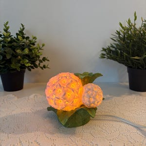 May include: A ceramic lamp shaped like two hydrangea flower clusters with a warm orange glow emanating from within. The flower petals are white, and the base consists of sculpted green leaves. A white power cord extends from the side of the base.
