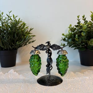 Peut inclure: Un ensemble décoratif de salière et poivrière. L'ensemble comprend deux salières en forme de grappe de raisin en verre vert avec des couvercles argentés, suspendues à un support en métal noir avec des détails de feuilles et de vigne. La base est ronde.