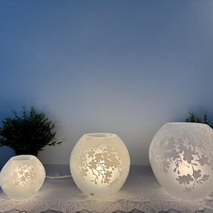 May include: Three frosted glass globe lamps with floral patterns, of different sizes, are lit on a white lace tablecloth. The lamps are arranged in a row, with a small green plant on either side.
