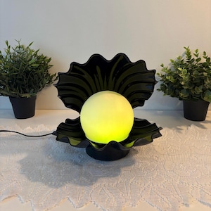 Peut inclure: Une lampe en forme de coquillage noir avec une ampoule sphérique lisse diffusant une lumière jaune au centre. La lampe présente un design à bords festonnés et comprend un fin cordon d'alimentation noir sortant de la base.