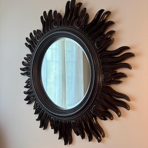 Peut inclure: Un miroir mural circulaire doté d'un cadre marron foncé avec un motif décoratif en forme de soleil. Le cadre se compose de multiples rayons incurvés en forme de flammes s'étendant vers l'extérieur, créant un motif radial sur le mur.