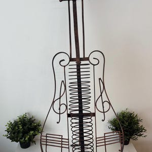 May include: A decorative metal music stand shaped like a cello. The stand is dark brown with a vertical spine and a series of horizontal lines and a central spring. Two small potted plants flank the stand.
