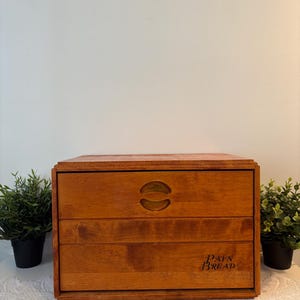 Puede incluir: Caja de pan de madera con un cálido acabado marrón rojizo. La caja tiene forma rectangular con tres cajones visibles. El cajón superior presenta un asa decorativa, y las palabras "PAIN BREAD" están impresas en el cajón inferior.