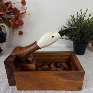 May include: A wooden nutcracker in the shape of a goose with a white head and a black beak. The nutcracker is open, revealing a wooden box filled with acorns. The goose's neck acts as the lever to crack nuts.