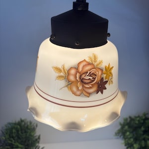 Könnte beinhalten: Vintage Pendelleuchte mit weißem Glasschirm mit floralem Design in Braun- und Gelbtönen. Der Schirm hat einen gewellten Rand und braune Streifen. Die Leuchte hat eine schwarze Metalloberseite.