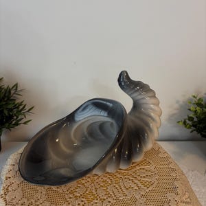 Peut inclure: Un bol décoratif en verre tourbillonnant gris et blanc en forme de corne d'abondance. Le bol a une large ouverture et une queue incurvée en forme de corne. Le bol est posé sur un napperon en dentelle crème.