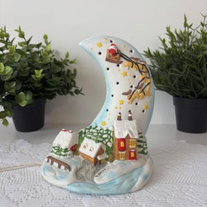 May include: A ceramic Christmas-themed night light shaped like a crescent moon. Santa Claus in a sleigh pulled by reindeer is depicted on the moon. The base features snow-covered houses and trees, with a light cord extending from the base.